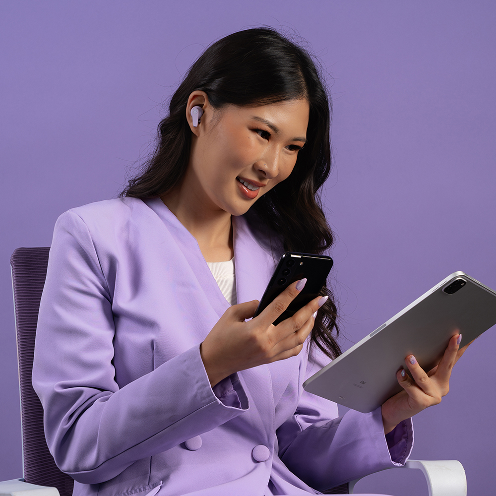 Kobieta w fioletowej marynarce używająca tableta i smartfonu mając założone słuchawki JLab Go Pods ANC.