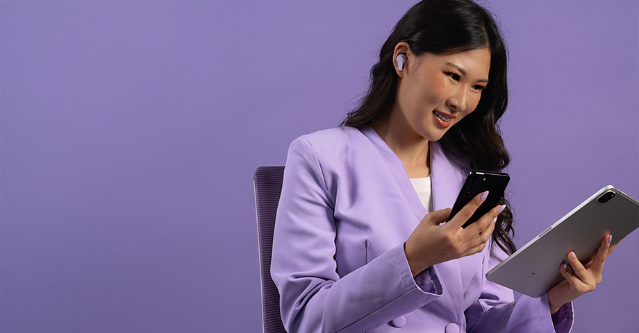 Kobieta w fioletowej marynarce używająca tableta i smartfonu mając założone słuchawki JLab Go Pods ANC.
