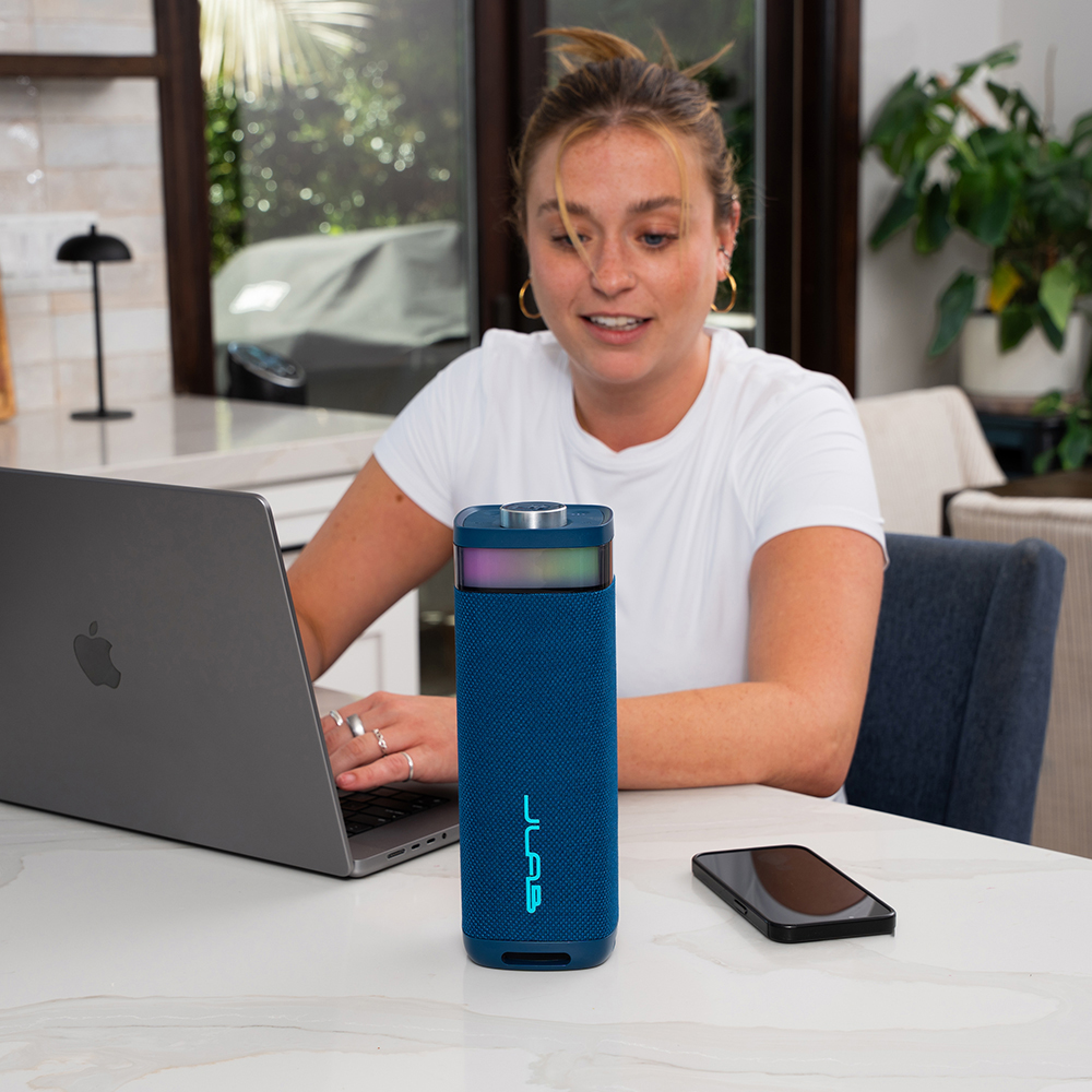 Niebieski JLab JBuds Party na stole obok laptopa i smartfonu.