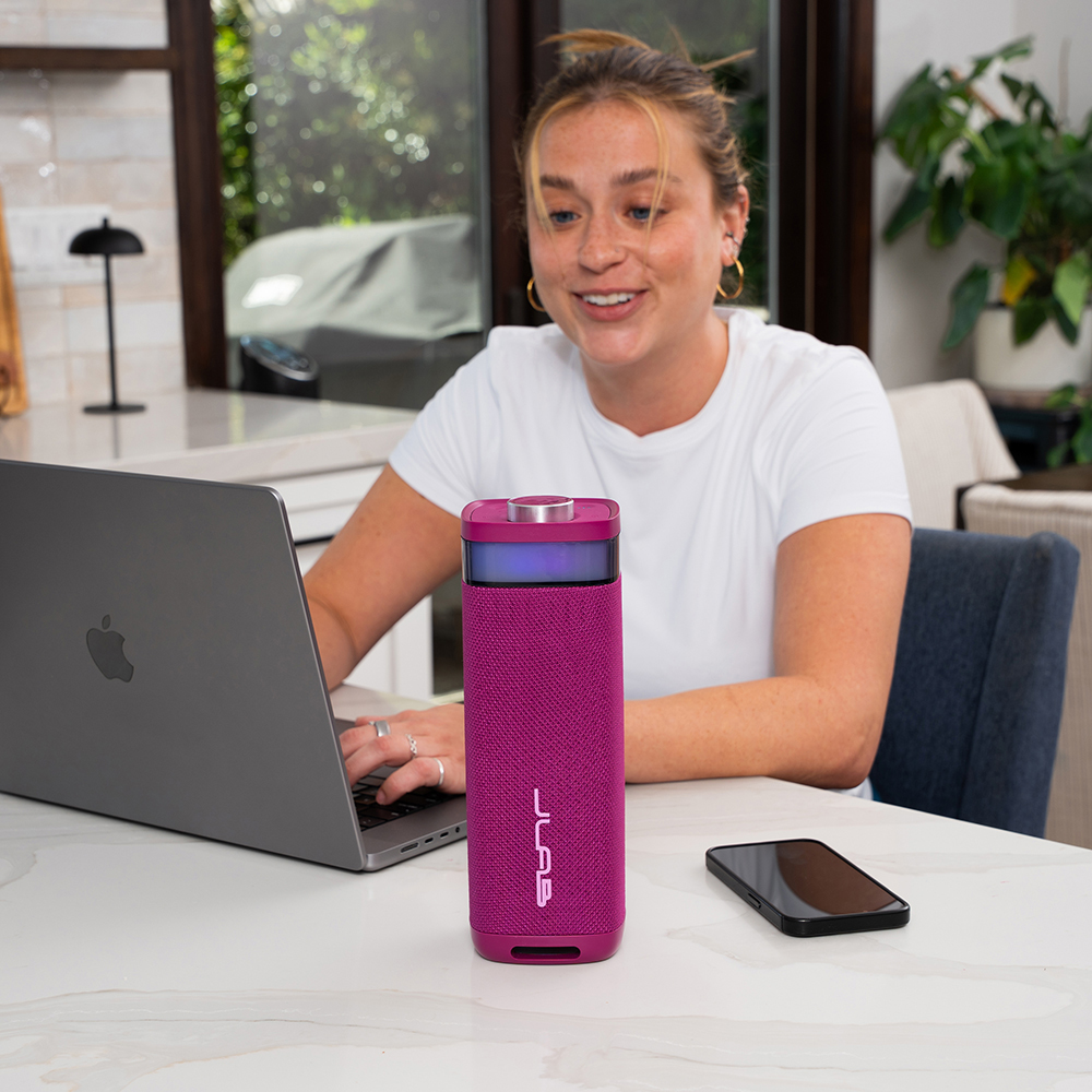 Różowy głośnik JLab JBuds Party na stole obok laptopa i smartfonu.