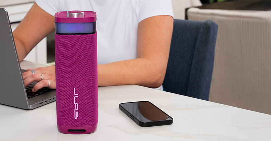 Różowy głośnik JLab JBuds Party na stole obok laptopa i smartfonu.