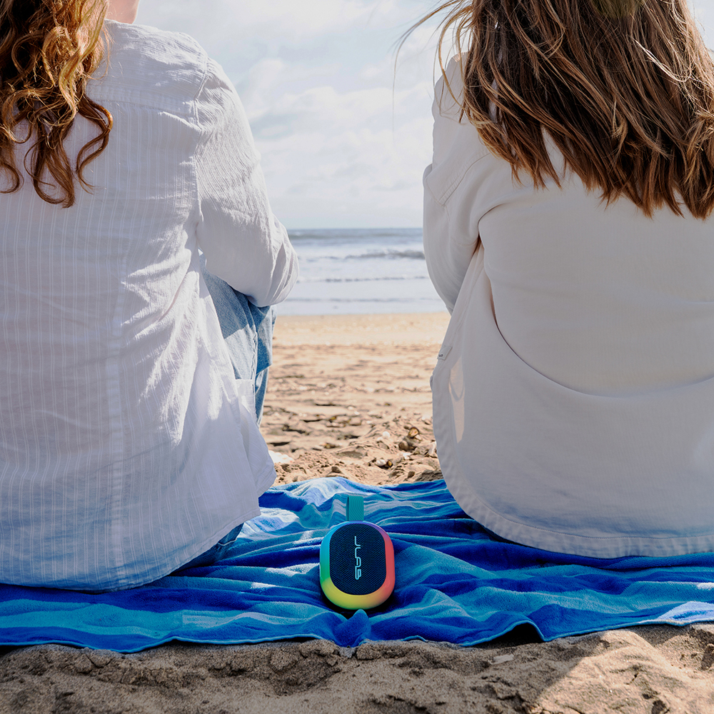 Dwie kobiety siedzące na kocu na plaży, pomiędzy nimi głośnik JLab Pop Party.