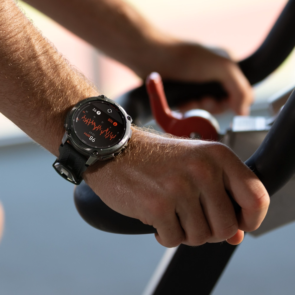 Zbliżenie na smartwatch Kospet Tank T4 mierzący tętno na nadgarstku mężczyzny trenującego na rowerze stacjonarnym.