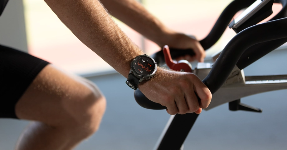 Smartwatch Kospet Tank T4 mierzący tętno na nadgarstku mężczyzny trenującego na rowerze stacjonarnym.