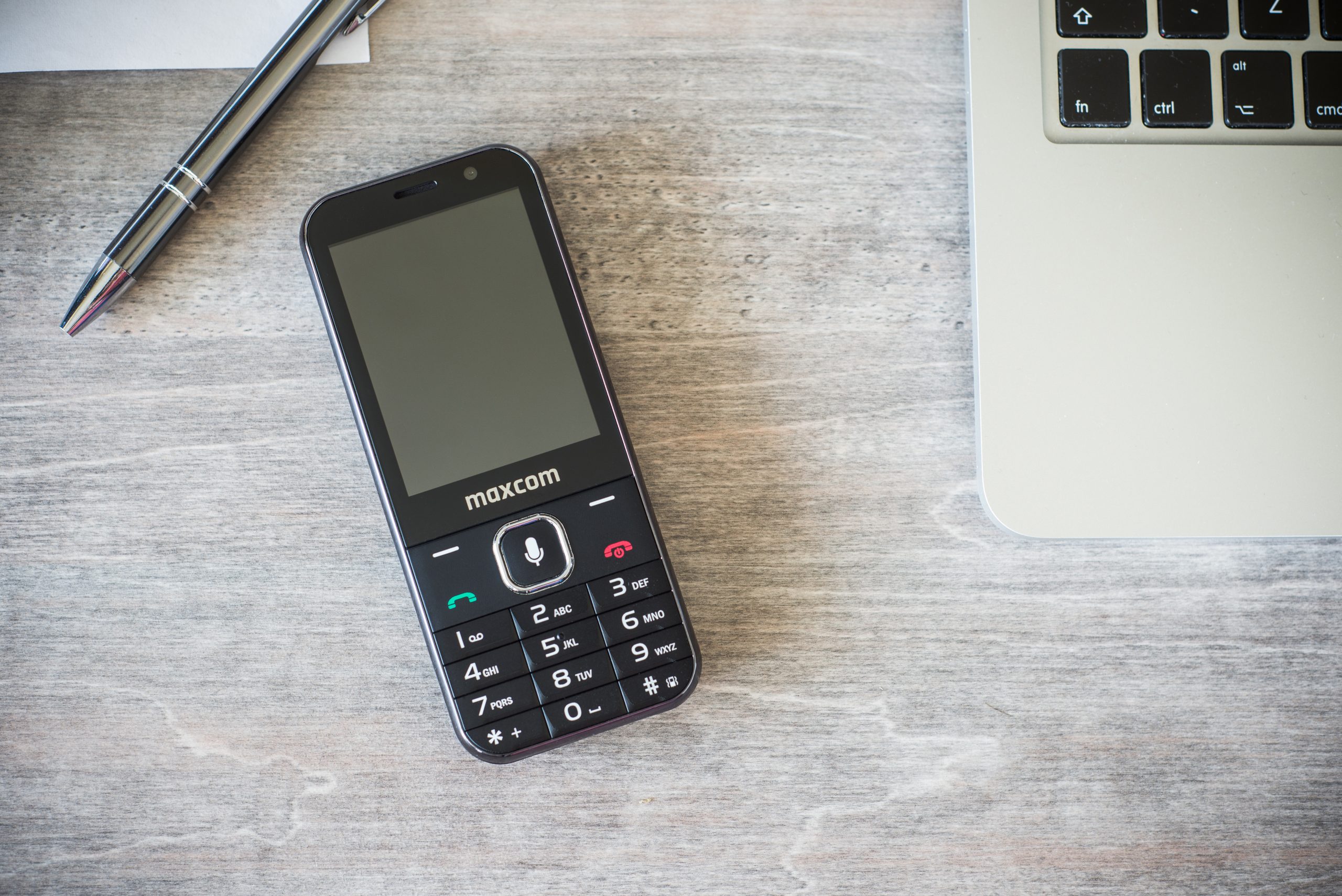 Telefon Maxcom z dużą klawiaturą leży na biurku obok laptopa i długopisu